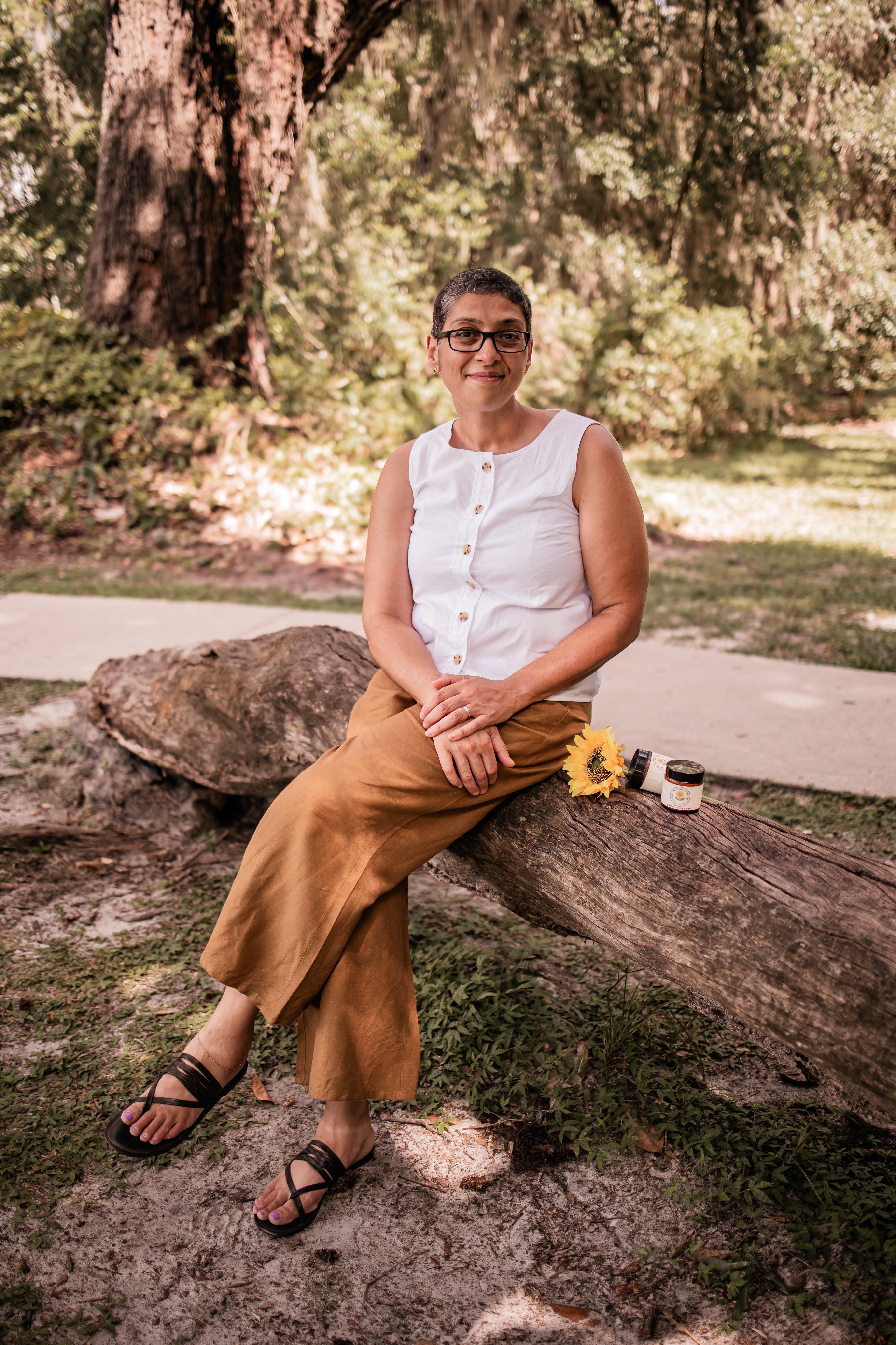 Chris Farlow, owner of Wildflower Glow, sitting on a log in a park-like setting with trees and grass.