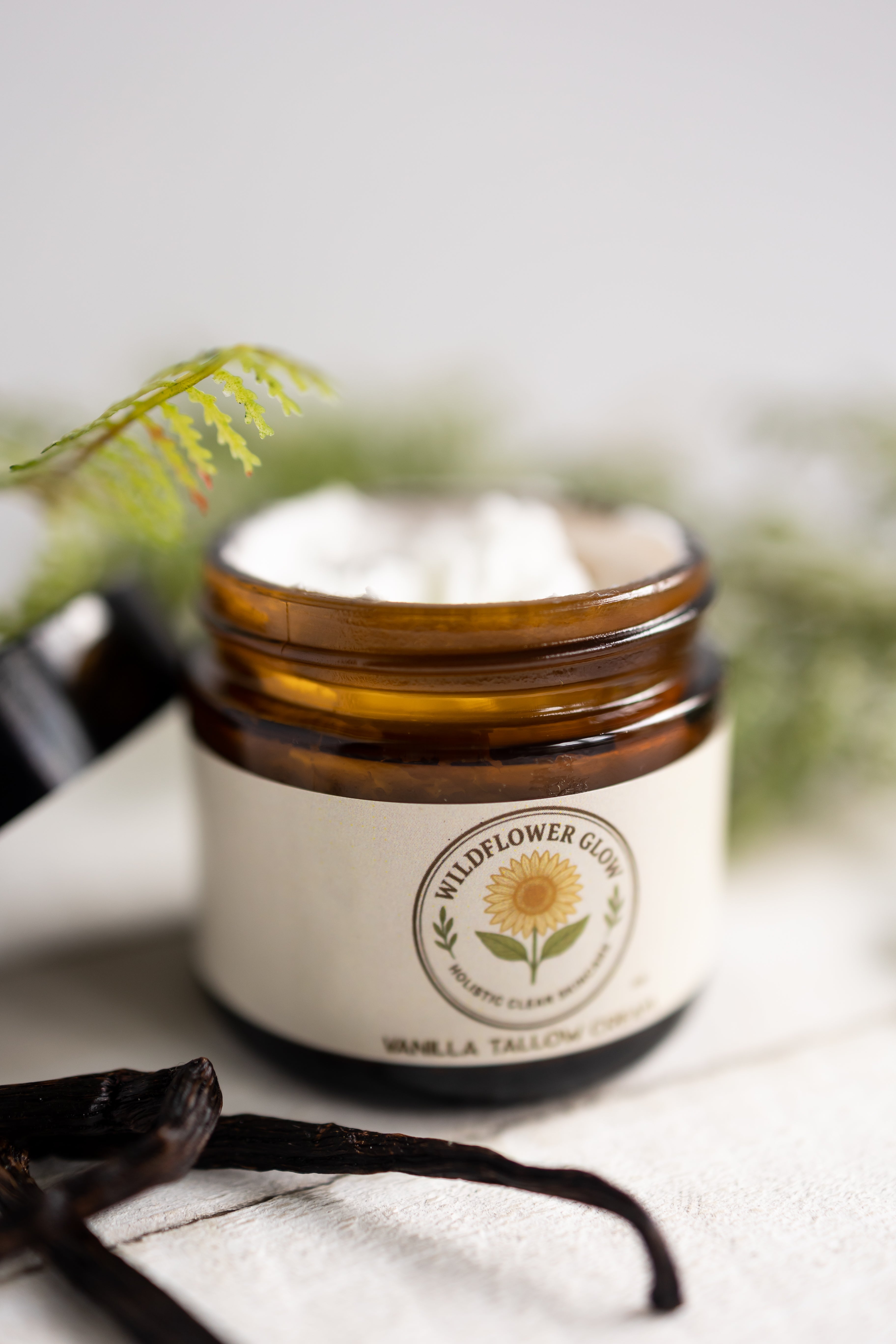 Jar of Wildflower Glow vanilla tallow cream with vanilla bean and leaves on a white surface