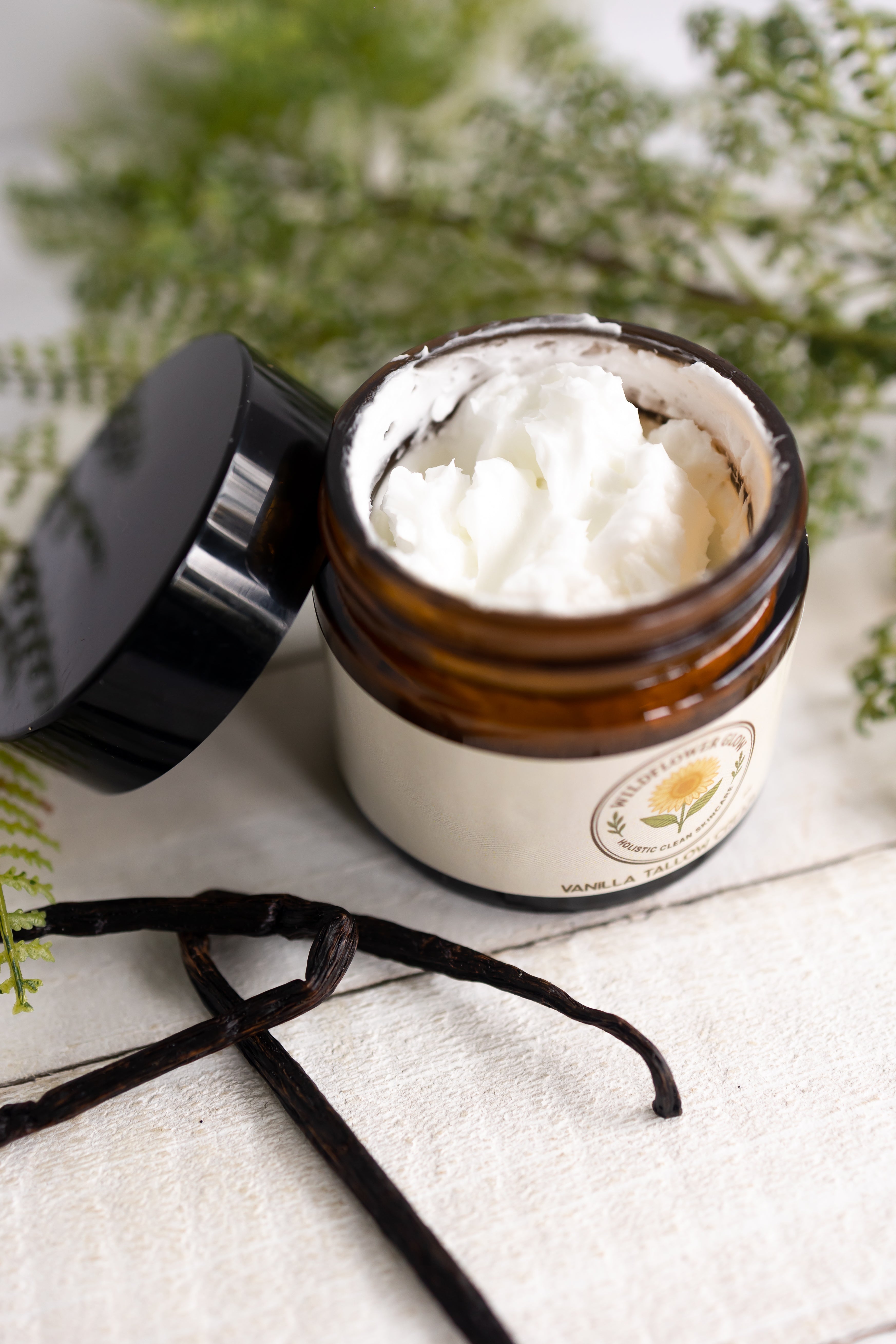 Jar of white cream with a vanilla bean on a light surface with greenery in the background