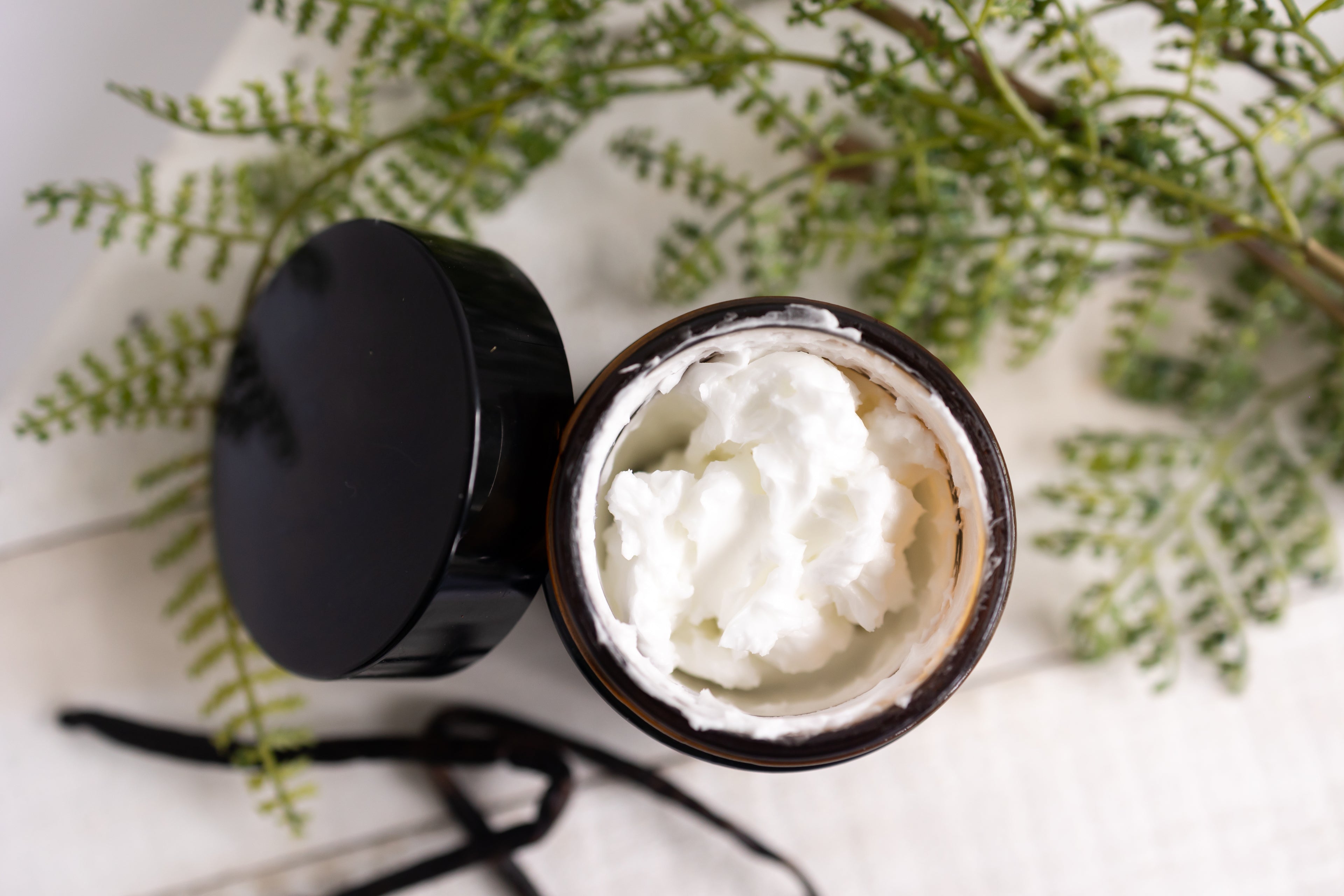 Jar of white cream with a black lid on a light surface with green leaves in the background