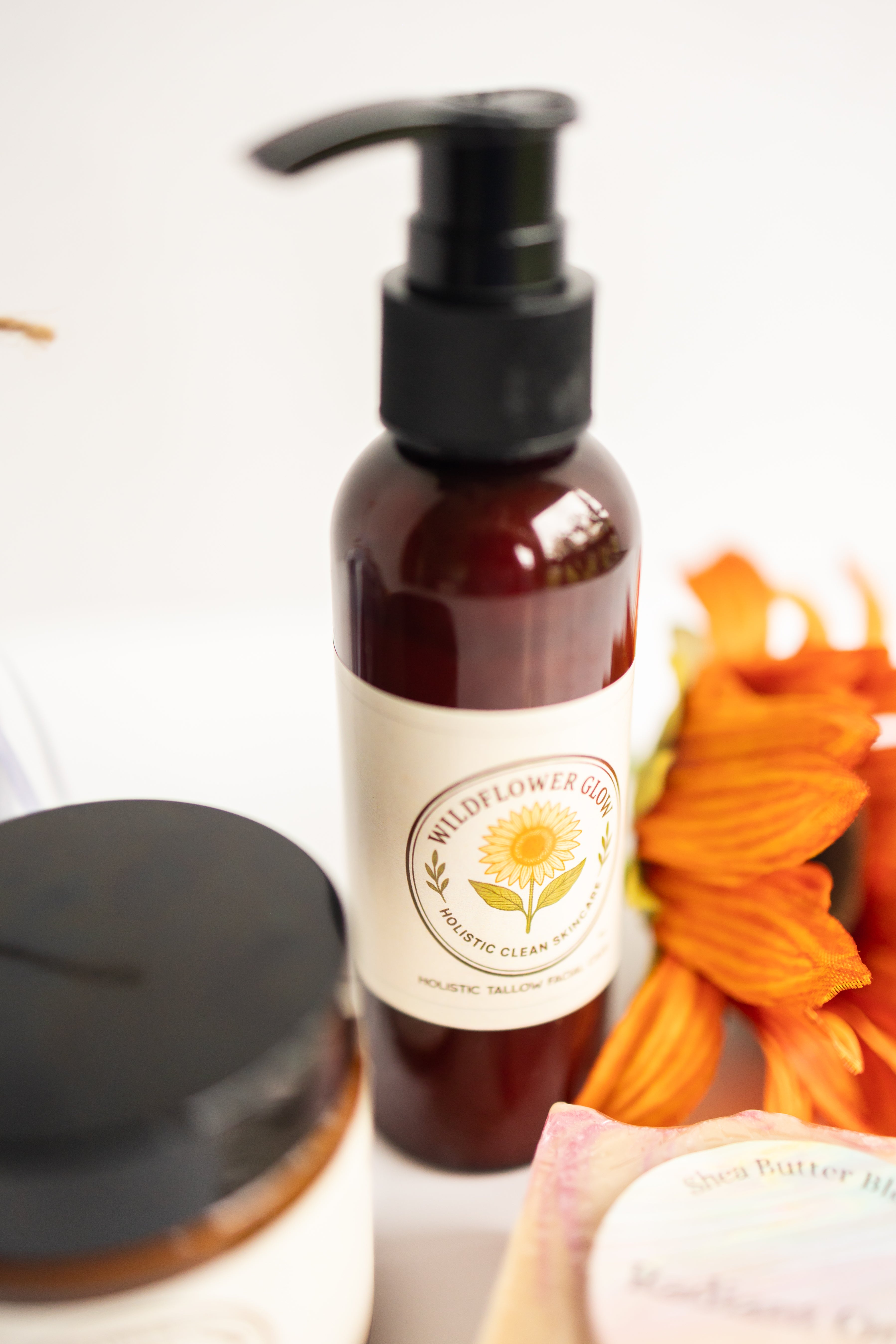 Bottle of Wildflower Glow Facial Cleanser with a label, surrounded by flowers on a light background