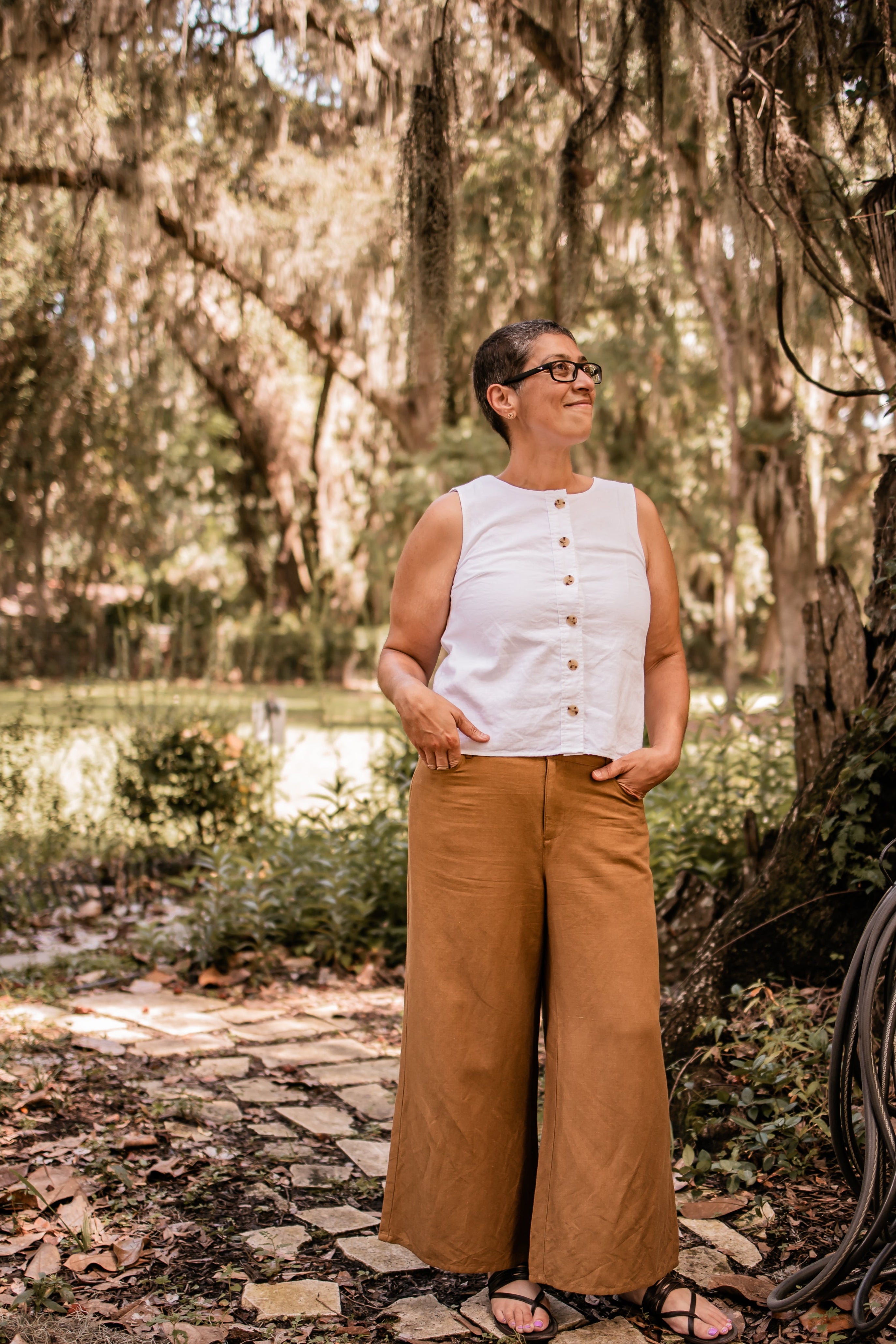 Chris Farlow, owner of Wildflower Glow, wearing a white sleeveless top and brown wide-leg pants standing in a natural setting with trees and a stone path.